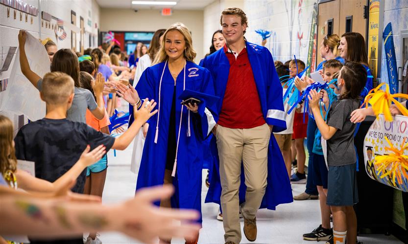 Students in graduation robes walk down an elementary hallway